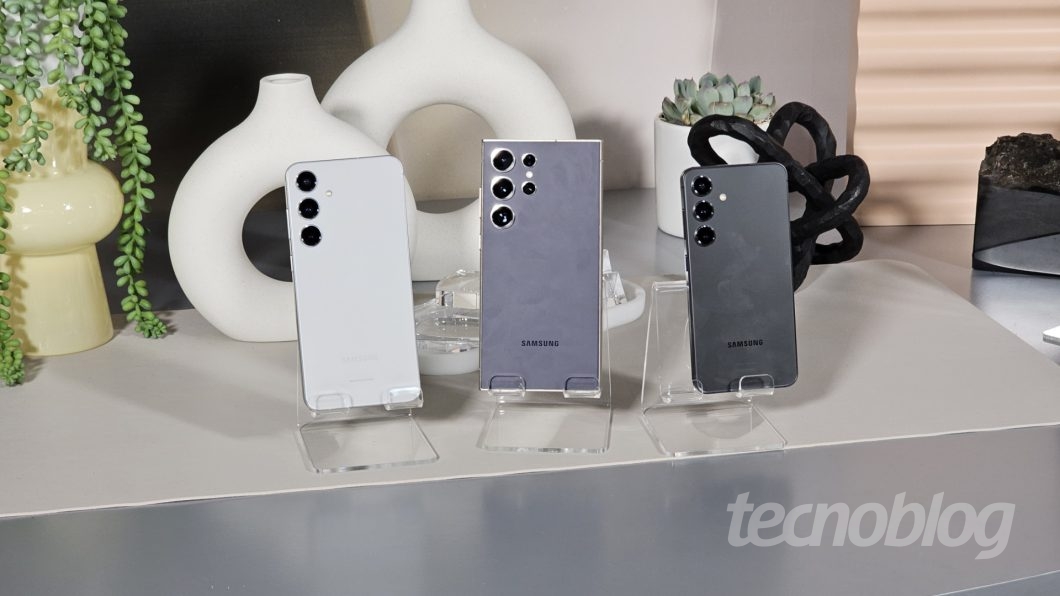 Três smartphones em suportes de acrílico sobre uma mesa com vasos e plantas decorativas