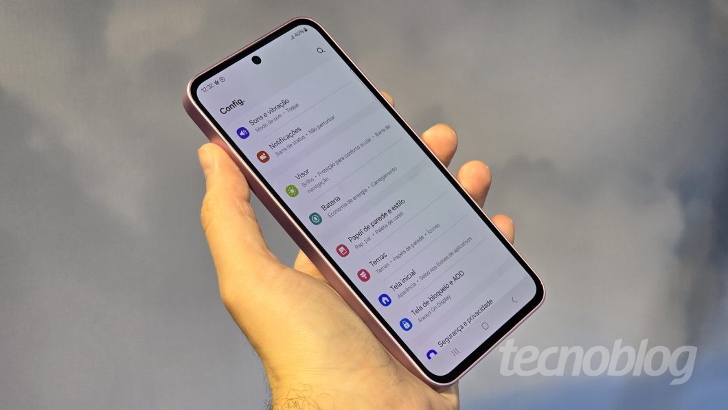 Pessoa segura um smartphone Samsung em tom rosado, com a tela exibindo o menu de configurações em português. São visíveis opções como "Sons e vibração", "Notificações", "Visor" e "Bateria". No canto inferior direito da imagem, há a marca d'água "tecnoblog". Fundo desfocado em tons cinzentos (foto: Thássius Veloso/Tecnoblog)