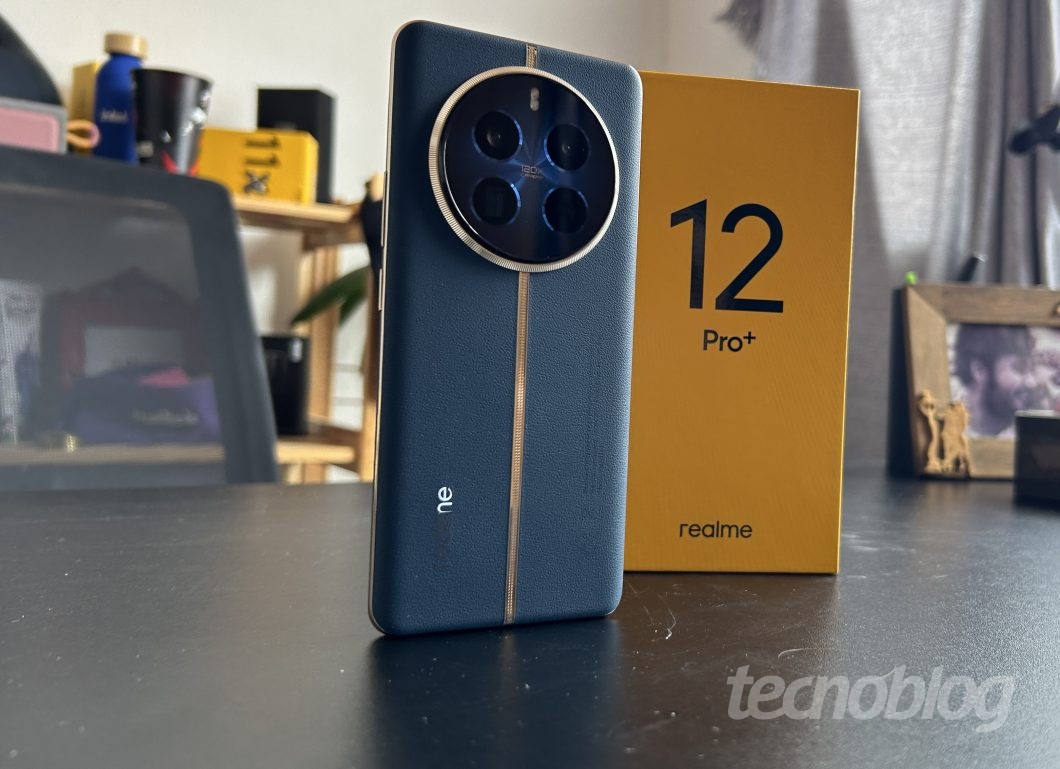 Celular Realme 12 Pro Plus na cor azul, em pé em uma mesa preta, em frente a caixa amarela do mesmo smartphone, em um cenário desfocado