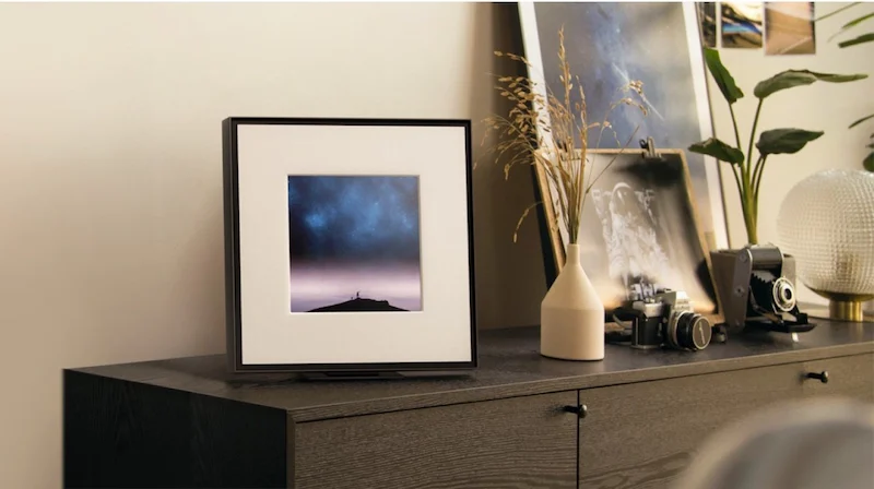 Alto-falante em formato de quadro de fotos sobre um móvel com flores e vasos.