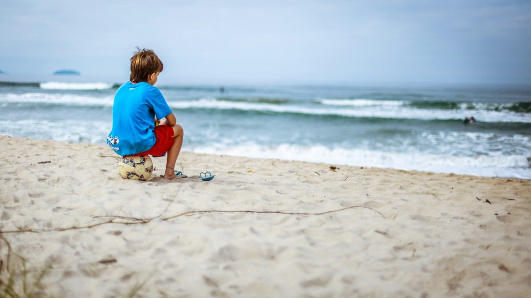 Criança sentada em uma bola de futebol, de frente para a praia