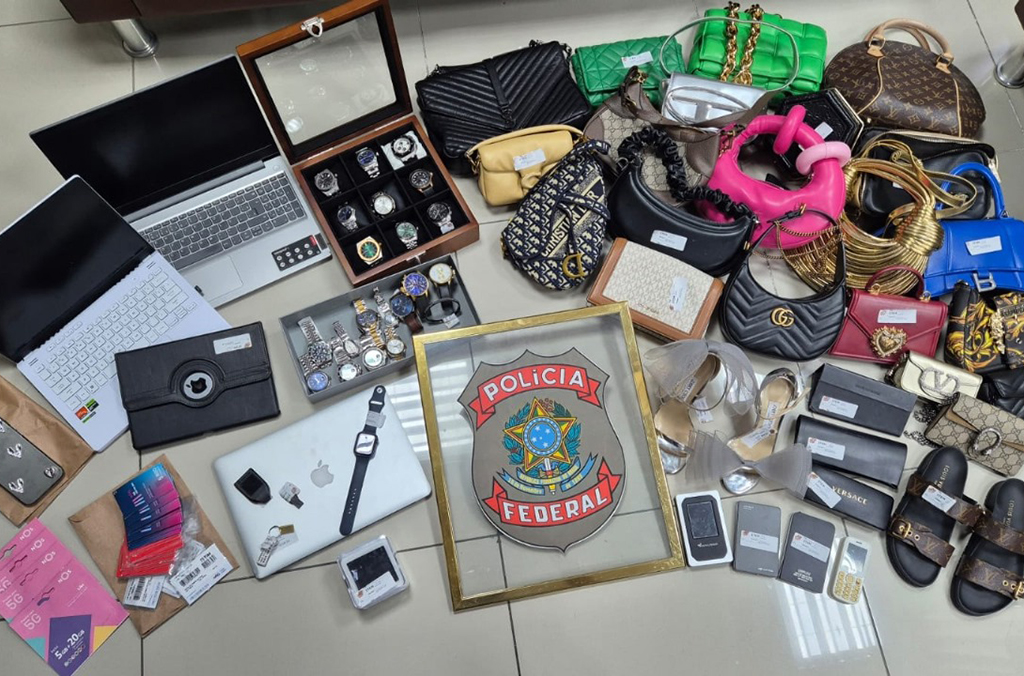 Notebooks, chips de telefonia, relógios, joias, bolsas e calçados no chão, junto à brasão da Polícia Federal