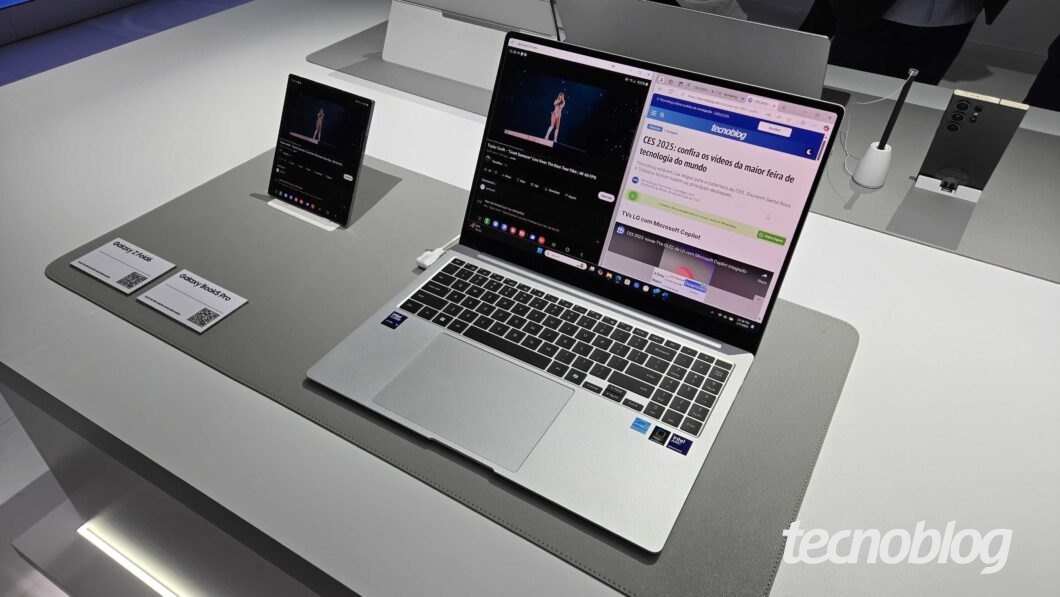 Notebook branco Galaxy Book 5 Pro está sobre uma mesa de exposição branca durante a CES 2025. A tela exibe ícones coloridos, com um show em reprodução numa metade e, na outra, o site do Tecnoblog.