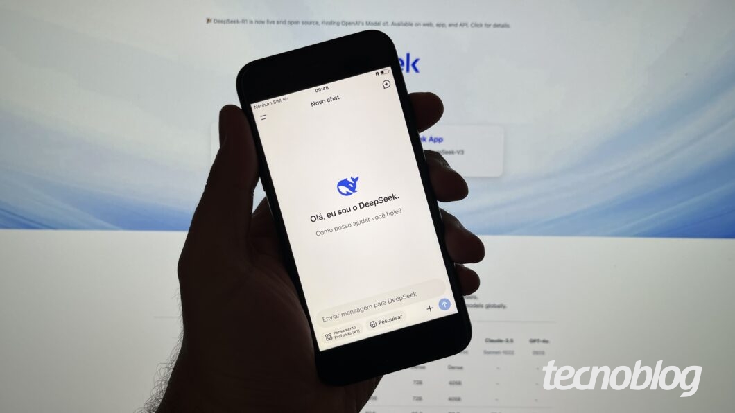 Mão segurando celular com o app do DeepSeek aberto. Ao fundo, o site do DeepSeek em um monitor.