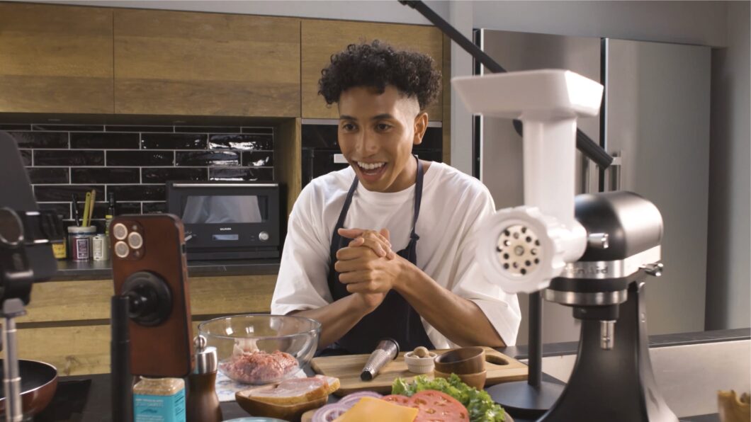 Homem em bancada de cozinha com alimentos a sua frente olhando para um celular em um tripé (Imagem: Divulgação)