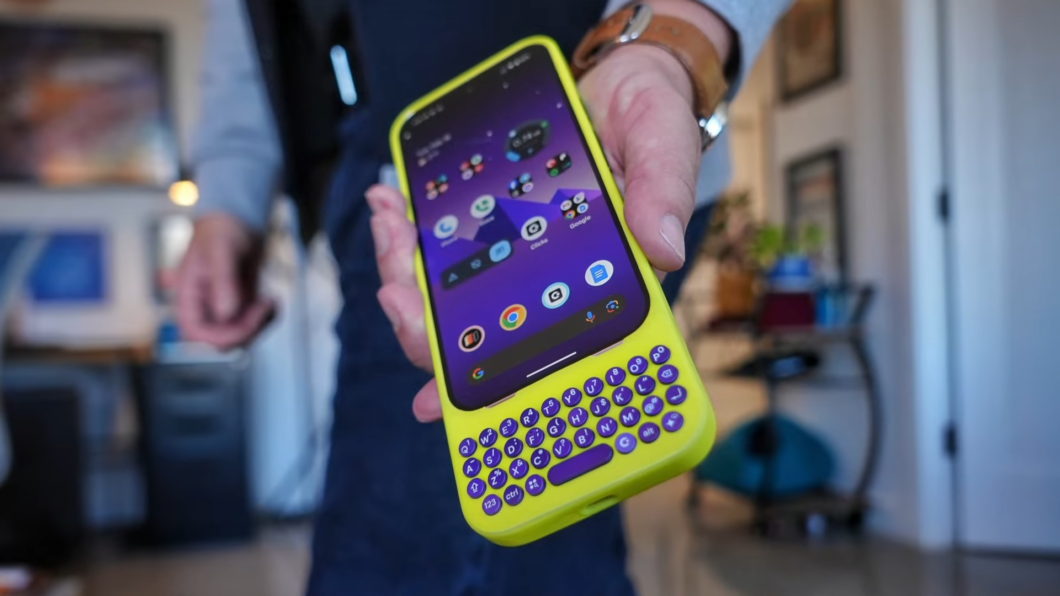 Um Google Pixel 9 em uma capinha Clicks amarela, que adiciona um teclado físico ao smartphone. A tela exibe a interface do Android com aplicativos organizados em pastas. A pessoa segura o aparelho com uma mão, mostrando o acessório em um ambiente interno.