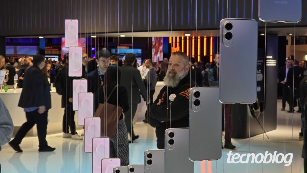 Fotografia mostra vários smartphones Galaxy S25 Edge exibidos em uma sala de exposição, pendurados por fios finos do teto e iluminados por baixo. À direita, celulares prateados são vistos de costas, revelando lentes de câmera duplas. À esquerda, smartphones com telas iluminadas em tons de rosa. Pessoas transitam ao redor dos displays, e o logotipo "tecnoblog" está no canto inferior direito.