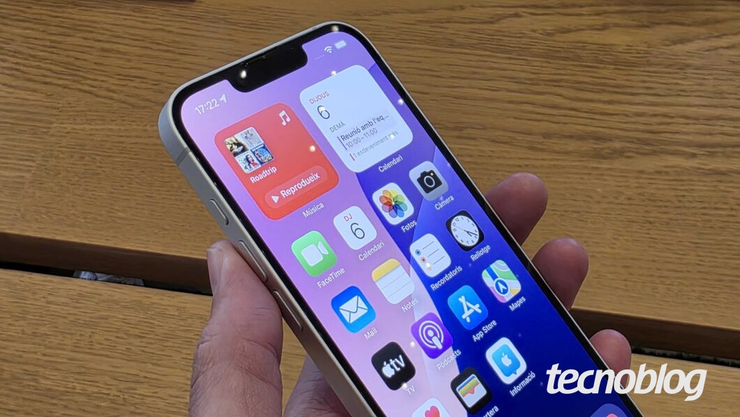 Imagem mostra um iPhone 16e branco sendo segurado em uma mão. Ele está com a tela desbloqueada, exibindo os aplicativos.