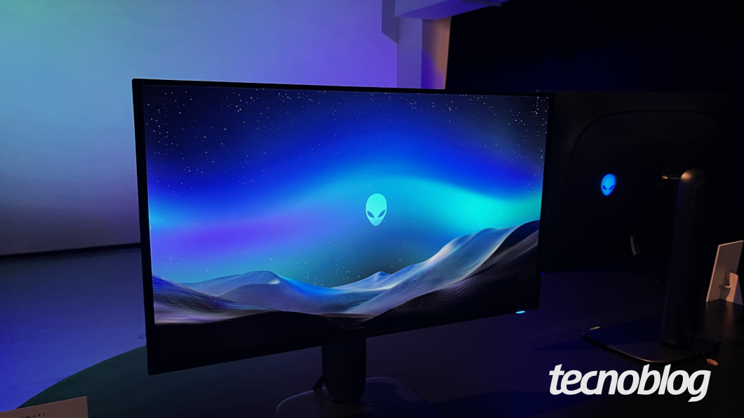 Monitor Alienware exibindo uma imagem com tema espacial, apresentando o logotipo da marca no centro da tela. O ambiente tem iluminação azulada e há outro monitor semelhante ao fundo.