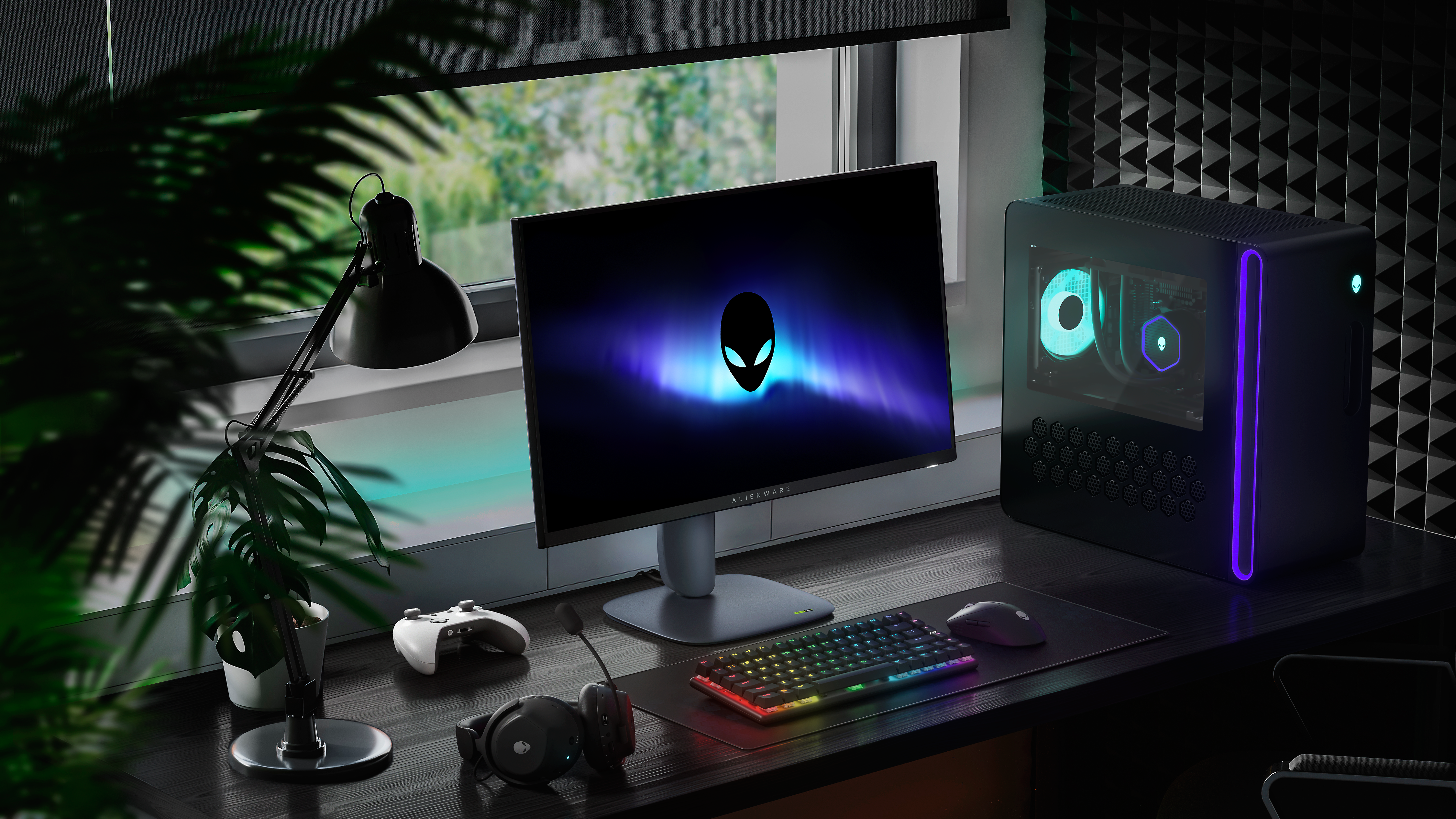 A imagem mostra um setup gamer moderno com um monitor Alienware exibindo um logotipo de alienígena, um PC com iluminação RGB e periféricos como teclado, mouse, headset e controle de videogame. A mesa é escura, e o ambiente tem iluminação LED, uma luminária de mesa e uma planta ao lado. Ao fundo, há uma janela com persiana e painéis acústicos na parede.