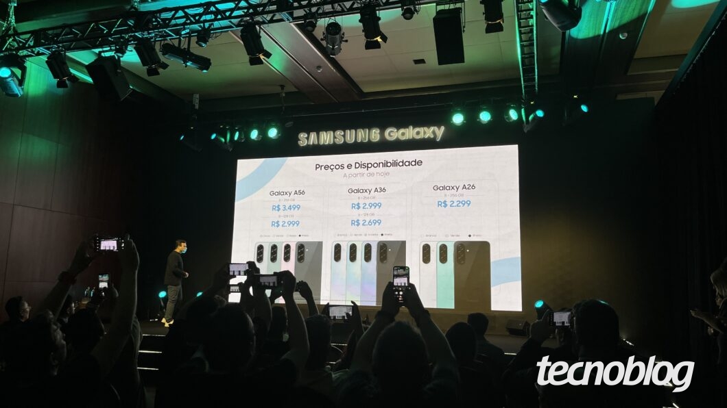 Imagem de um evento de lançamento da Samsung para os smartphones Galaxy A56, A36 e A26 5G. O telão mostra os seguintes preços: Galaxy A56 8GB + 256GB por R$ 3.499 e 8GB + 128GB por R$ 2.999. Galaxy A36 8GB + 256GB por R$ 2.999 e 8GB + 128GB por R$ 2.699. Galaxy A26 8GB + 256GB por R$ 2.299. Os dispositivos estão disponíveis nas cores branco, verde e preto.