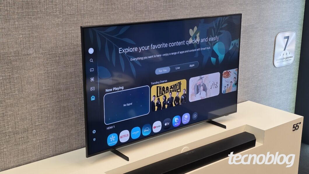 Tela de smart TV de 55 polegadas exibindo menu inicial com opções de apps como Netflix, Prime Video, Disney+, YouTube e Apple TV. No centro da tela, texto em inglês: "Explore your favorite content quickly and easily" e abaixo, seleção de dramas em destaque. A TV está sobre suporte branco e há uma soundbar preta à frente. Ao lado, placa com texto "7 anos Atualização garantida Sistema Operacional Tizen".