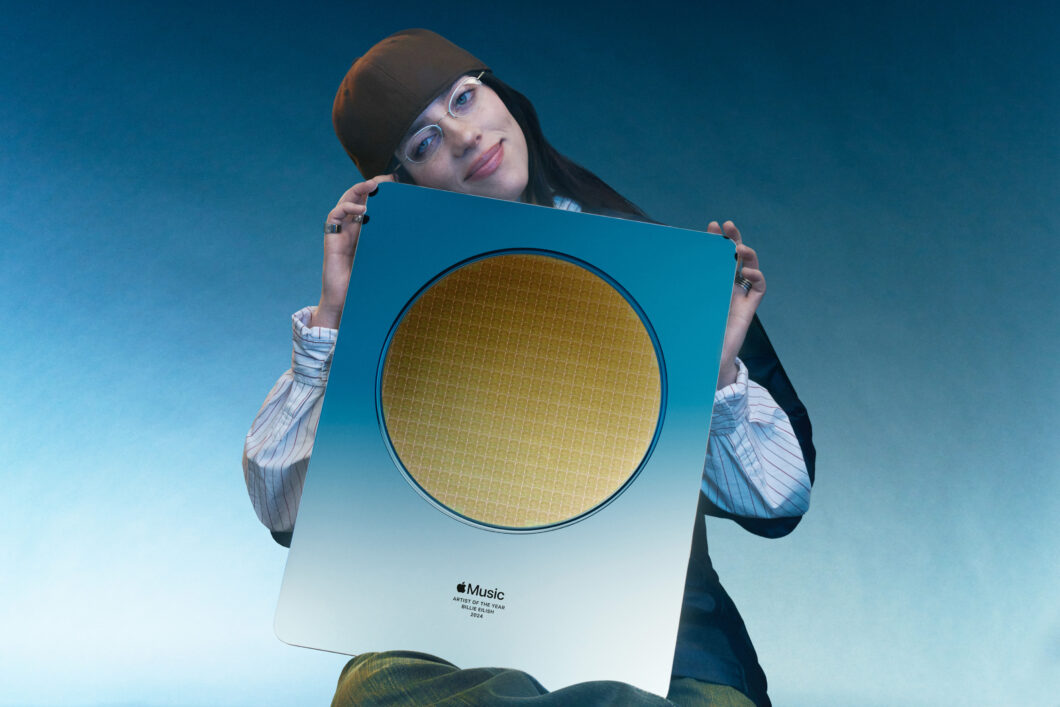 A cantora Billie Eilish, usando óculos e gorro marrom, segura um prêmio metálico com um disco dourado no centro. Na parte inferior, lê-se: "Apple Music Artist of the Year 2024". Fundo em gradiente azul.