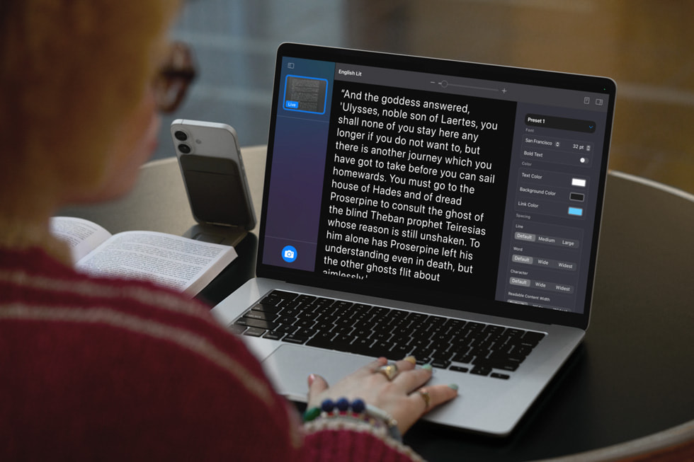 Uma pessoa com óculos e um suéter vermelho está sentada a uma mesa preta, usando um Macbook aberto que exibe um aplicativo de leitura ou anotação de texto, com um zoom que deixa as letras grandes. O texto na tela parece ser um trecho literário em inglês, com formatação e opções de personalização visíveis em um painel lateral direito, incluindo configurações de fonte, tamanho, cor do texto e fundo. À esquerda do laptop, um smartphone está apoiado em um suporte, e um livro aberto está ao lado.