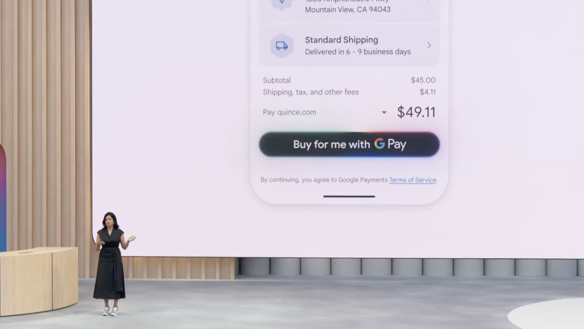 Mulher em palco apresentando nova função de pagamento Google Pay em evento do Google. Tela grande atrás mostra endereço, opção de frete padrão com prazo de entrega de 6 a 9 dias úteis, subtotal de , taxa de ,11 e botão "Buy for me with G Pay". Cenário com elementos em madeira clara e logotipo "G" do Google.