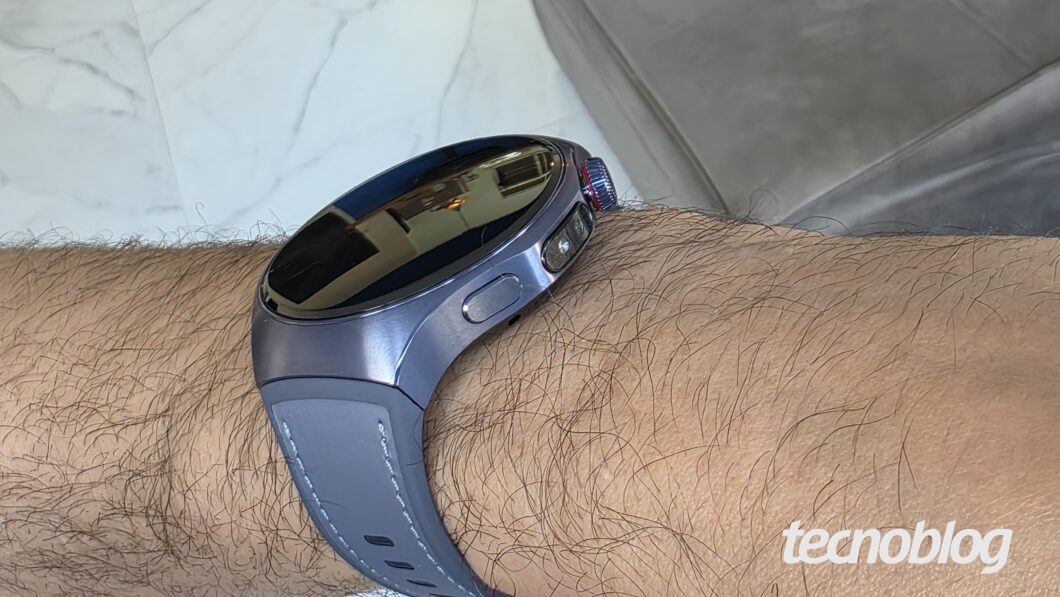 Imagem em close mostra um smartwatch com caixa metálica prateada e pulseira azul clara, usado em um pulso masculino. A tela do relógio está desligada e reflete parcialmente o ambiente ao redor. O fundo apresenta piso branco e parte de um sofá cinza. O relógio tem dois botões laterais, um deles com detalhes em vermelho.