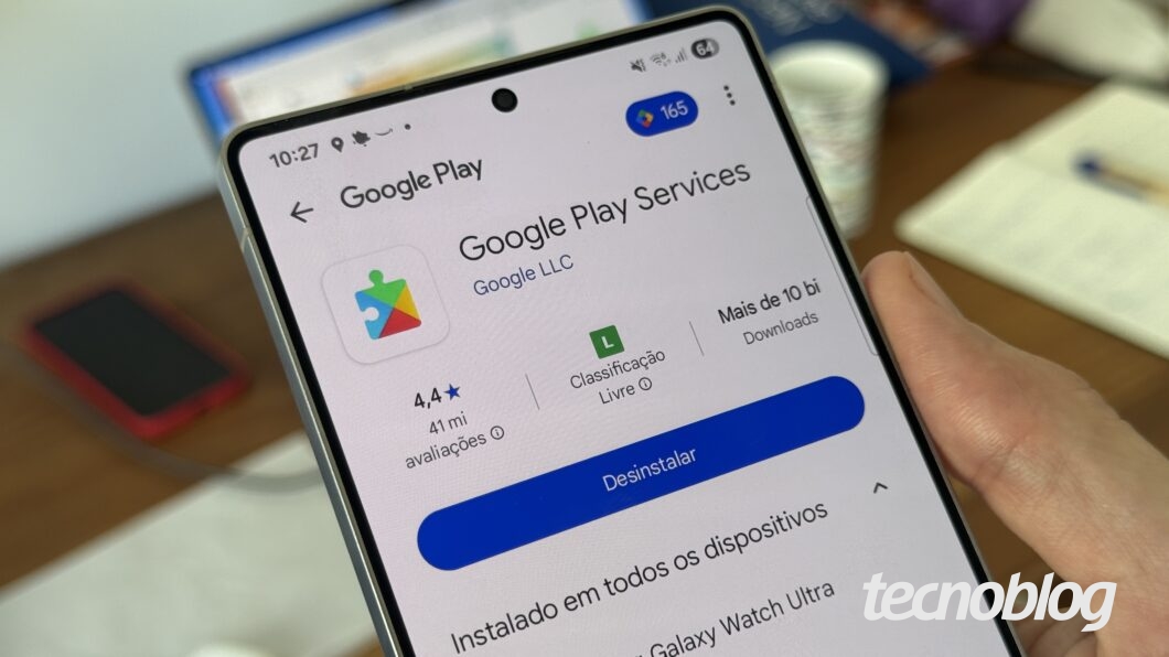 Tela de um smartphone exibindo a página do aplicativo "Google Play Services" na Google Play Store. A avaliação média é de 4,4 estrelas com 41 milhões de avaliações, classificação livre e mais de 10 bilhões de downloads. O botão azul exibe "Desinstalar". Ao fundo, um smartphone vermelho, um notebook e papéis sobre uma mesa.