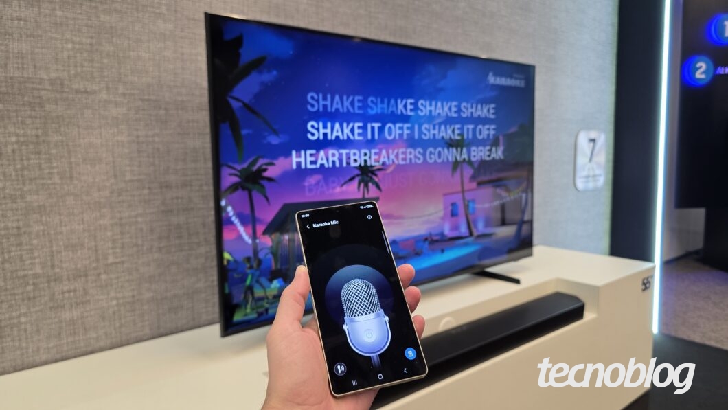 Mão segura smartphone com app de microfone para karaokê aberto, TV ao fundo exibe letras da música "Shake it off".