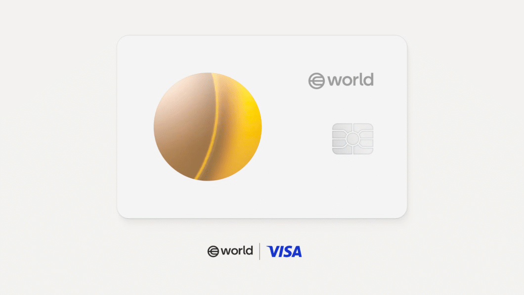 Uma imagem de um cartão de crédito branco com um círculo dividido verticalmente em tons de bege e amarelo na lateral esquerda. À direita do círculo, a palavra "@world" está escrita em letras pequenas e cinzas. Abaixo, um chip de cartão de crédito prateado é visível. Na parte inferior central, as palavras "@world | VISA" aparecem em letras cinzas. O fundo é branco.