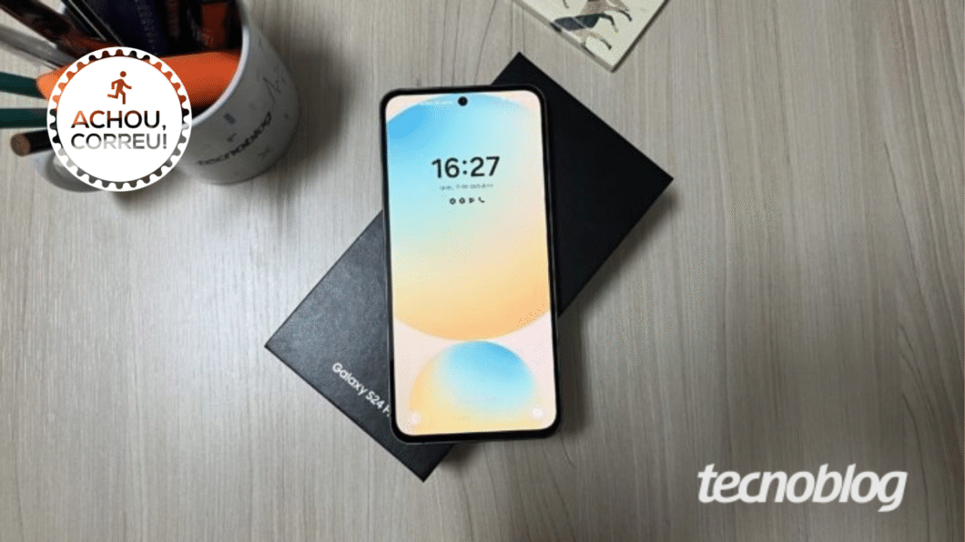A imagem mostra um Celular Samsung Galaxy S24 FE ligado, com a tela virada para cima, em cima de uma caixa preta sobre uma mesa de madeira