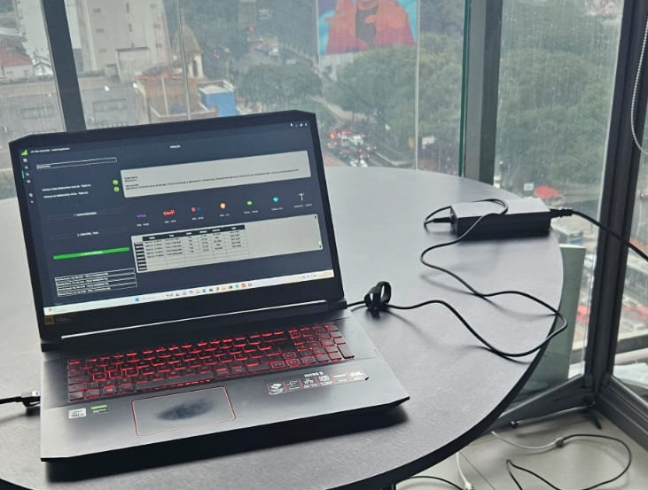 Laptop com teclado iluminado em vermelho sobre mesa redonda preta, com carregador conectado. Tela mostra interface de software com gráficos e tabelas. Ambiente interno, próximo a janela de vidro com vista para edifício e rua movimentada ao fundo, em dia nublado (imagem: divulgação)