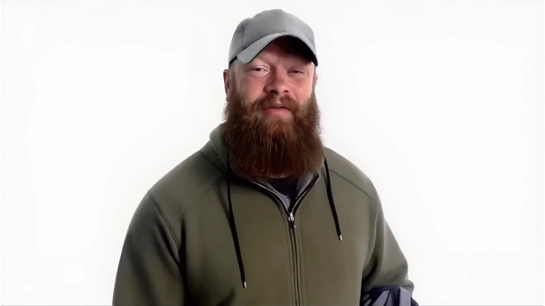 Homem de barba ruiva espessa e longa, vestindo um boné cinza e um agasalho verde-oliva com zíper, posa diante de um fundo branco. Ele está com uma expressão leve, de olhos parcialmente fechados, e parece estar em um ambiente de estúdio iluminado