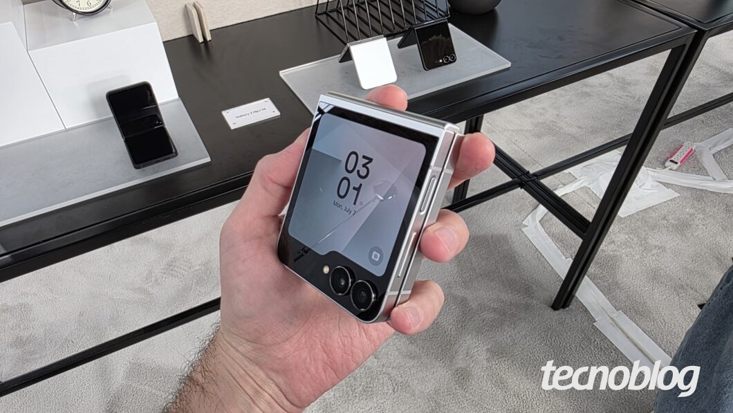 Mão segurando smartphone dobrável fechado, com tela externa acesa indicando 03:01h