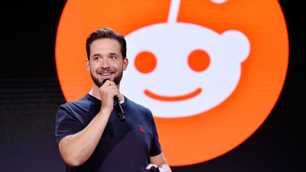 Imagem mostra um homem de camiseta preta, segurando um microfone, e o logo do Reddit ao fundo, em cor laranja