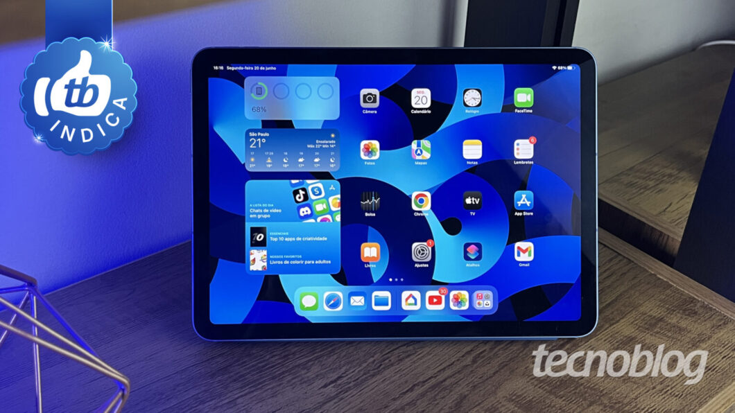 Tablet Apple iPad mostrando a tela inicial com apps e widgets, em uma mesa com iluminação azul