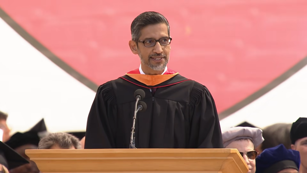 O executivo Sundar Pichai, homem de pele retinta clara, óculos e barba grisalha aparada, fala ao microfone atrás de um púlpito de madeira. Ele veste uma beca de formatura preta com detalhes em laranja e vermelho nos ombros.