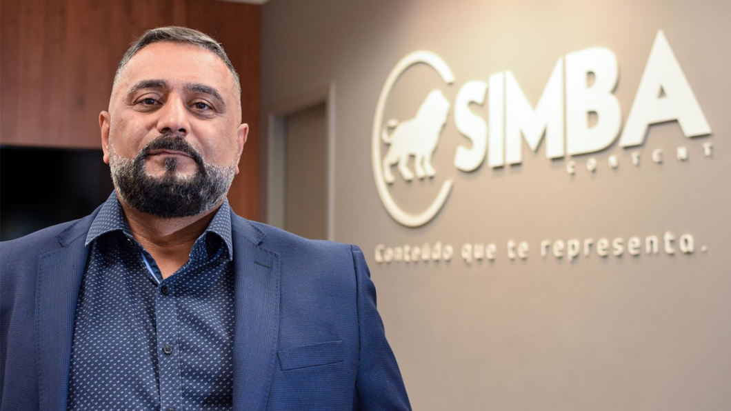 Foto de Carlos Alkimim, de terno. Ele posa em ambiente corporativo, com o logotipo da Simba Content ao fundo e a frase “Conteúdo que te representa” na parede.