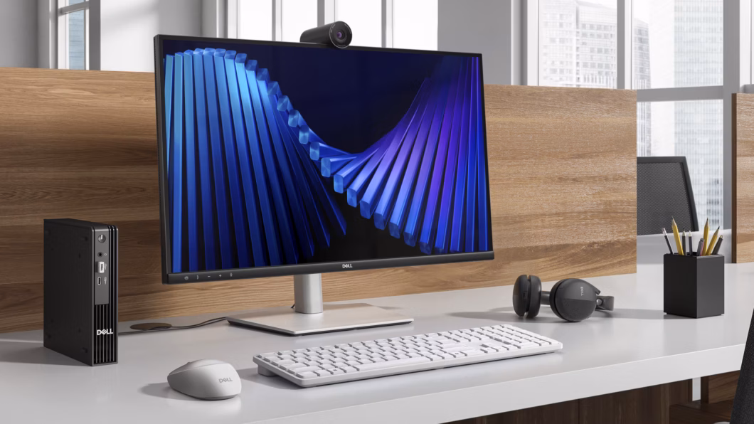 Mesa de escritório com monitor Dell, webcam, mini PC, teclado, mouse e headset, compondo uma estação de trabalho corporativa.