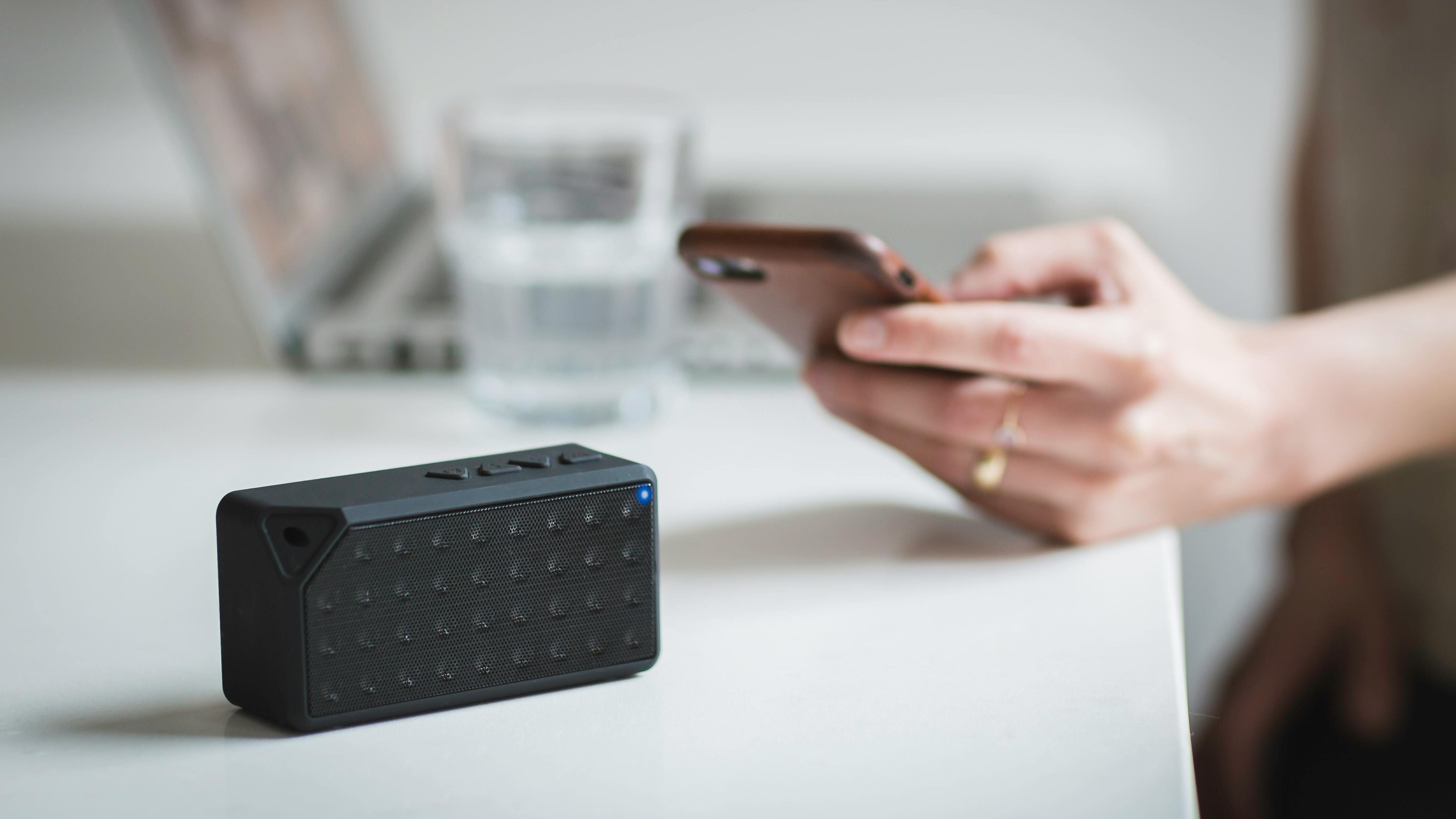 Alto-falante Bluetooth sobre mesa, com mão segurando smartphone próxima. Ao fundo um laptop e um copo d'água