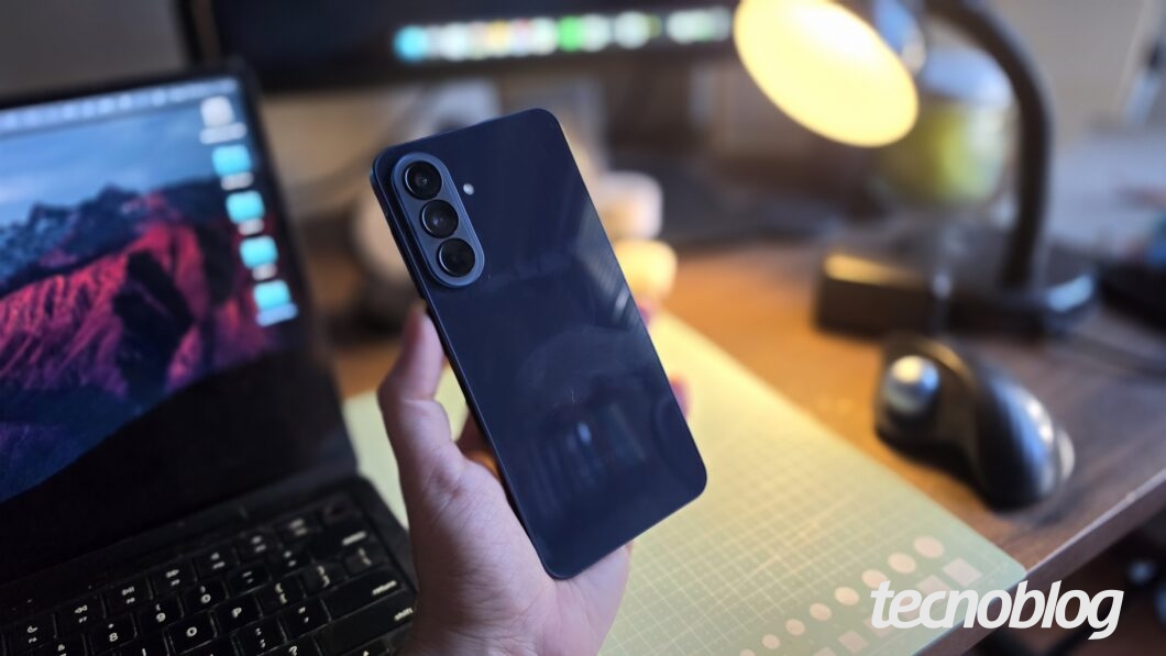 Pessoa segurando um smartphone azul com câmera traseira, em uma mesa de trabalho com notebook ao fundo