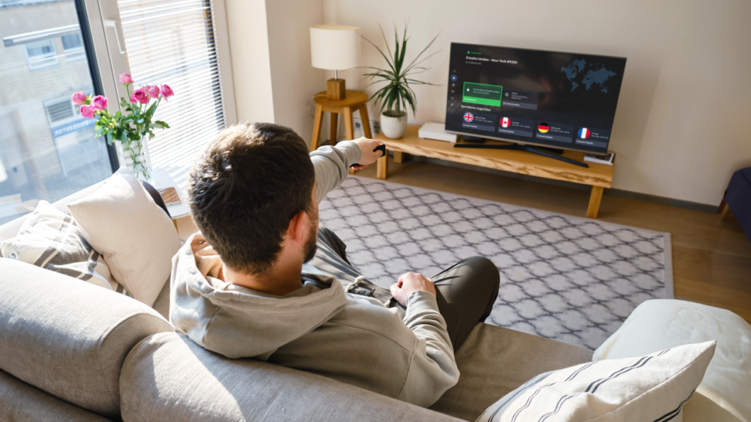 Pessoa sentada no sofá usa uma TV com a NordVPN aberta, exibindo opções de conexão e servidores em diferentes países.