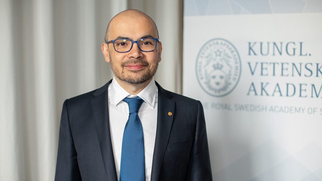 Foto de Demis Hassabis ao lado de uma placa branca. Ele veste um blazer preto, com uma gravata azul e uma camisa branca.