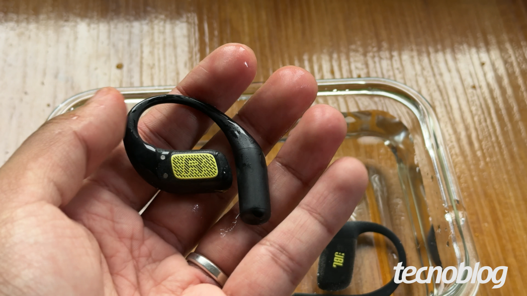 Mão segurando o JBL Endurance Zone molhado em teste, com a água ao fundo (resistência IP68)