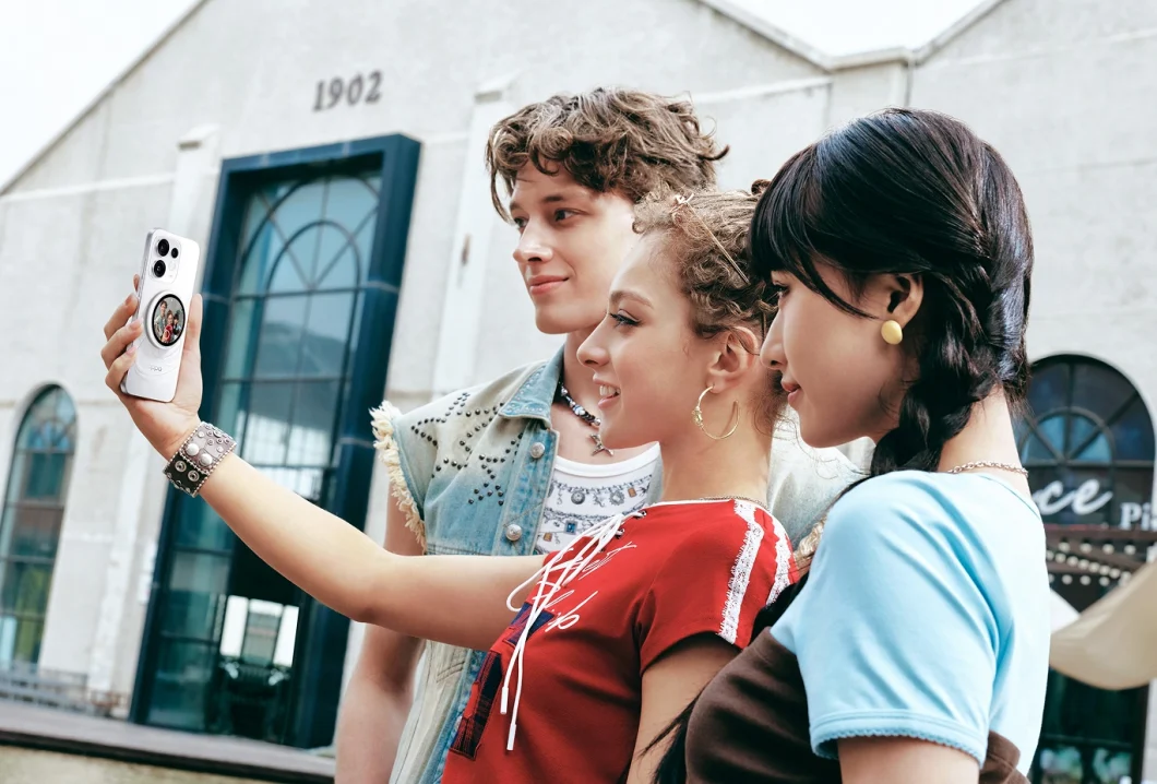 Três jovens posam para uma selfie ao ar livre usando o OPPO Reno16 branco, com o smartphone em destaque na mão de uma delas.
