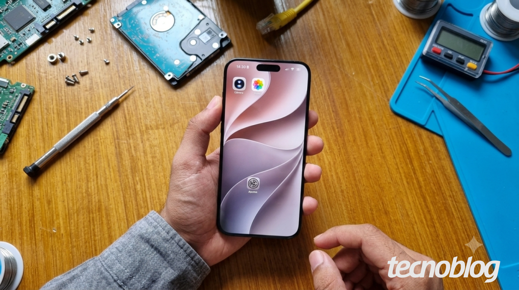 Mão segura um iPhone em uma bancada com componentes eletrônicos, ilustrando que a versão beta pode afetar desempenho e bateria
