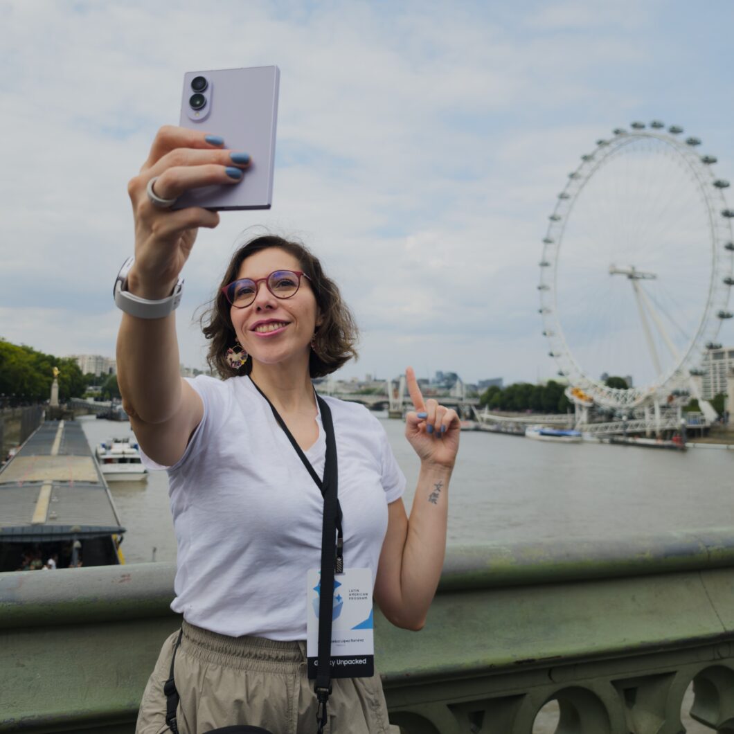 Mulher sorrindo tirando uma selfie com celular em frente à roda-gigante ao fundo