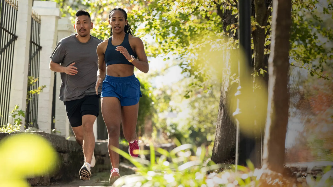 Um casal de pessoas correndo ao ar livre, passando por um jardim com árvores e luz do sol. A mulher utiliza um Garmin Forerunner 570 no pulso.