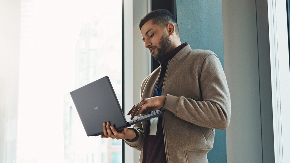 Homem de casaco bege e crachá segurando notebook Dell aberto com uma mão e digitando com a outra, próximo a uma janela ampla em escritório.