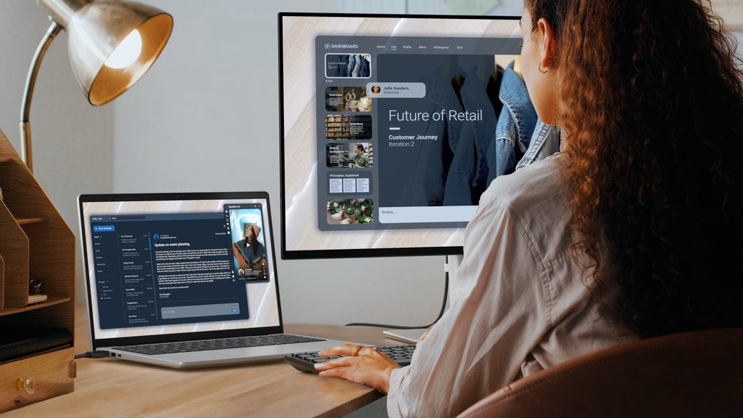 Mulher de costas sentada em mesa de trabalho utilizando notebook conectado a um monitor externo grande, que exibe apresentação de slides com o título