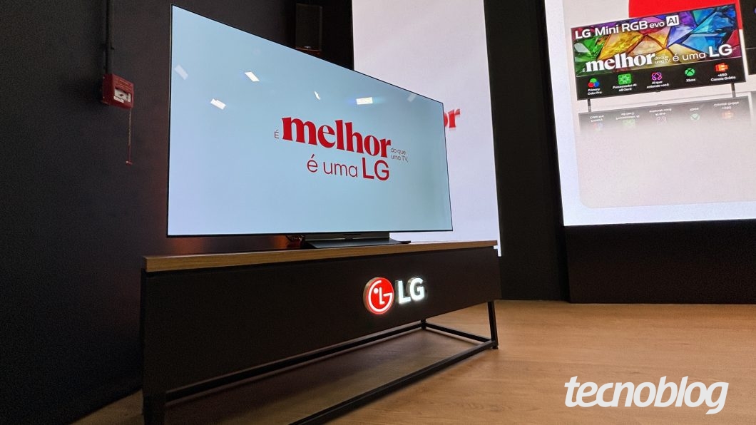 Smart TV ligada e apoiada sobre um rack escuro com tampo de madeira clara, em um ambiente de exposição. Na frente do móvel, destaca-se a logomarca iluminada da "LG". A tela branca do televisor exibe centralizado o texto em vermelho "É melhor do que uma TV, é uma LG". No canto esquerdo, na parede ao fundo, há um caixote de alarme vermelho. À direita, observa-se parte de outro painel publicitário com imagens e legendas sobre o modelo "LG Mini RGB evo AI".