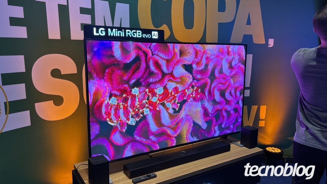 Smart TV de tela grande posicionada sobre um móvel de madeira clara. No topo da TV, há uma placa iluminada com a inscrição "LG Mini RGB evo AI". A tela exibe uma imagem colorida com textura de flores rosa e vermelhas e joias encrustadas. À frente do painel, sobre o móvel, estão uma soundbar preta e um controle remoto. Ladeando a soundbar, há duas caixas de som pretas. No lado direito, vê-se a parte posterior do corpo de uma pessoa vestindo camiseta cinza.
