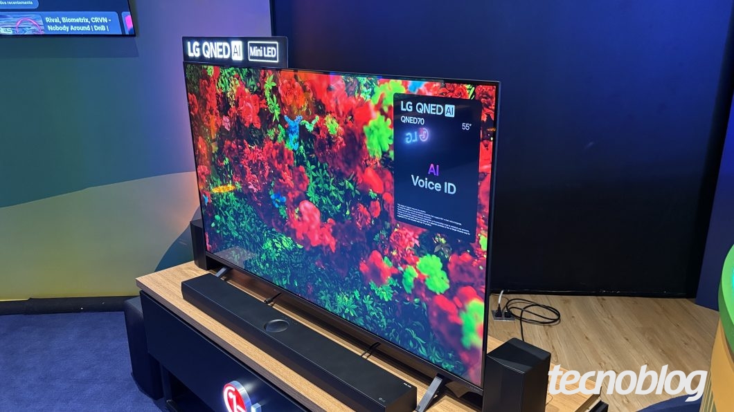 Televisor de tela plana exibido em ângulo diagonal sobre um móvel de madeira. Na parte superior do painel, uma placa preta traz os dizeres "LG QNED AI Mini LED". A tela exibe uma imagem vívida com folhagens verdes e flores vermelhas. No canto superior direito do display, sobressai um quadro preto informativo com os textos "LG QNED AI", "QNED70", "55"" e "AI Voice ID". Uma soundbar preta está posicionada logo à frente da base da TV, acompanhada por caixas de som laterais.