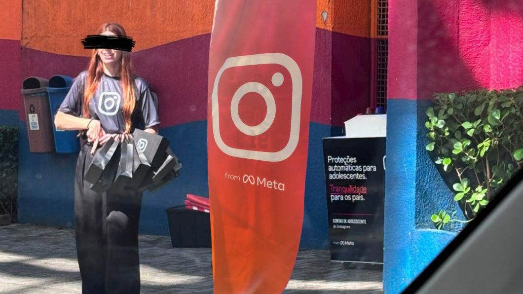 * Fotografia em ambiente externo. À esquerda, uma mulher de cabelos ruivos longos e tarja preta nos olhos veste camiseta cinza com o ícone do Instagram e segura sacolas pretas. Ao centro, destaca-se uma bandeira vertical laranja com o ícone do Instagram em branco e a frase