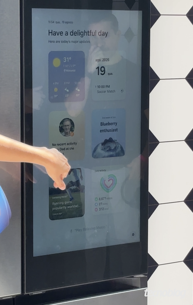 Uma mão de pele clara aponta para a tela de uma geladeira inteligente. A interface exibe o texto "Have a delightful day", seguido de dados em blocos. O primeiro mostra a temperatura "31°" em "Morumbi". Ao lado, a data "19 ago. 2026" e "10:00 PM Soccer Match". Abaixo, um card tem a foto de um homem e o texto "No recent activity from Dad". Outro exibe uma mirtilo com o título "Blueberry enthusiast". Na parte inferior, há uma notícia com um corredor e um indicador de "Daily activity" com "8,671 steps".
