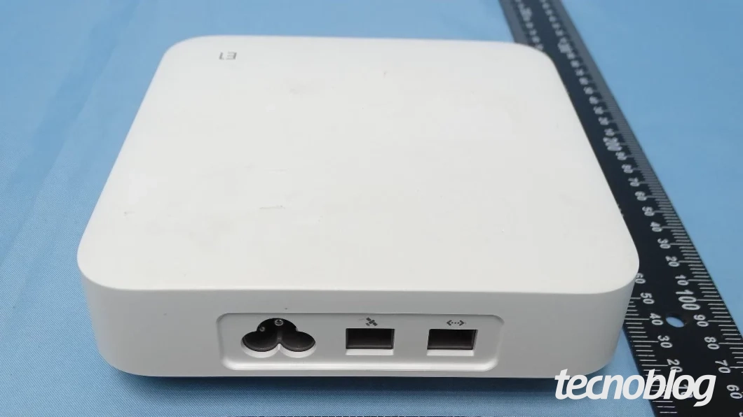 Fotografia técnica de homologação de um roteador de cor branca. Ele exibe a gravação cinza "L3" na parte superior esquerda de sua superfície e as portas de conectividade; o aparelho está apoiado sobre uma mesa azul e posicionado ao lado de uma régua milimetrada preta para medição de suas dimensões, trazendo o logotipo do "tecnoblog" impresso em branco no canto inferior direito.