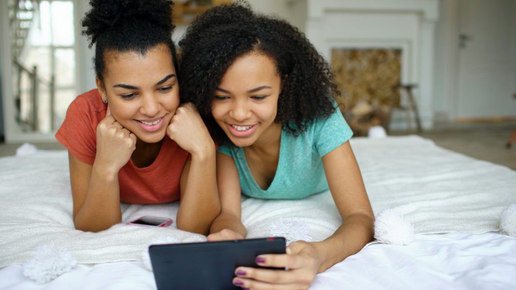 Duas adolescentes sorrindo e usando um tablet na cama, enquanto conversam e acessam conteúdo juntos